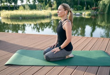 Daha iyi ol. Sportif orta yaşlı kadın rahat görünüyor, gözleri kapalı oturuyor meditasyon yaparken, yeşil bir parkta yoga yapıyor.