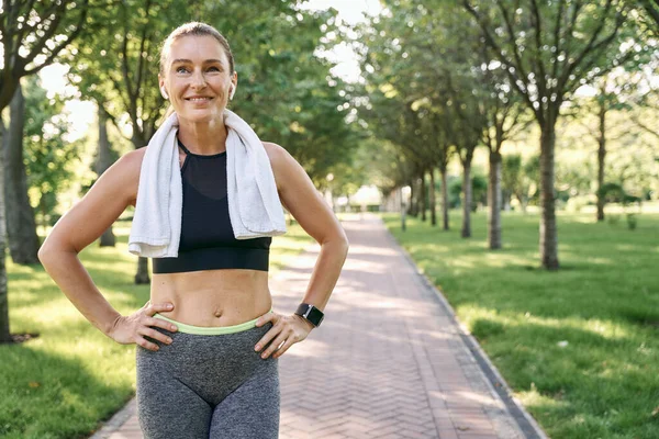 Stay active. Happy sporty woman in sportswear and wireless earphones smiling away, standing with towel around her neck after jogging in a green park on a sunny day