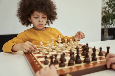 Saçı afro saçlı şirin çocuk satranç oynarken hareket etmeden önce düşünüyor, masada oturuyor.