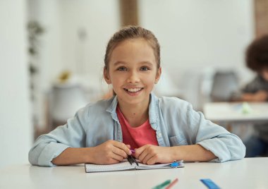 Güzel öğrenci. Sınıftaki masada otururken kameraya gülümseyen güzel küçük okul kızı.
