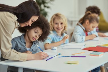 İlköğretim sınıfında diğer çocuklarla aynı masada otururken bayan öğretmeni dinleyen küçük özenli öğrenci.