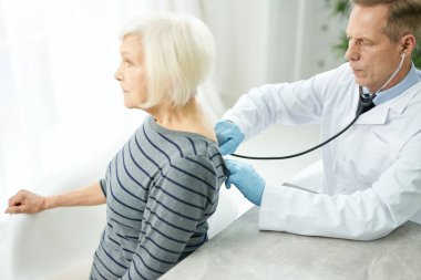 Steteskopla klinikte yaşlı kadını muayene eden ciddi bir doktor.