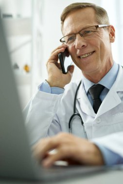 Cep telefonuyla konuşan ve klinikte dizüstü bilgisayar kullanan neşeli bir doktor.