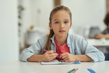Küçük tatlı liseli kız sınıftaki masada oturmuş kameraya bakıyor.