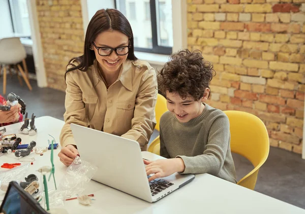 Learning to code. Smiling young female teacher in glasses showing scientific robotics video to little boy, using laptop in classroom