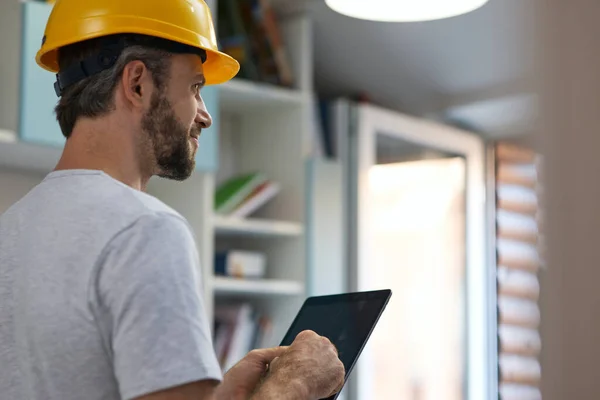 Reparasi profesional memakai helm sambil melihat ke samping, menggunakan tablet digital ketika berdiri di dalam ruangan — Stok Foto