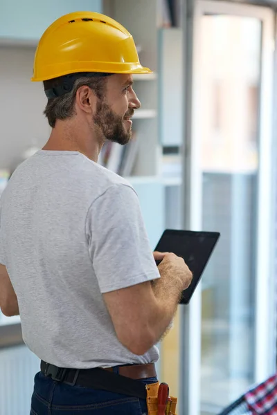 Reparasi profesional memakai helm sambil melihat ke samping, menggunakan tablet digital ketika berdiri di dalam ruangan — Stok Foto