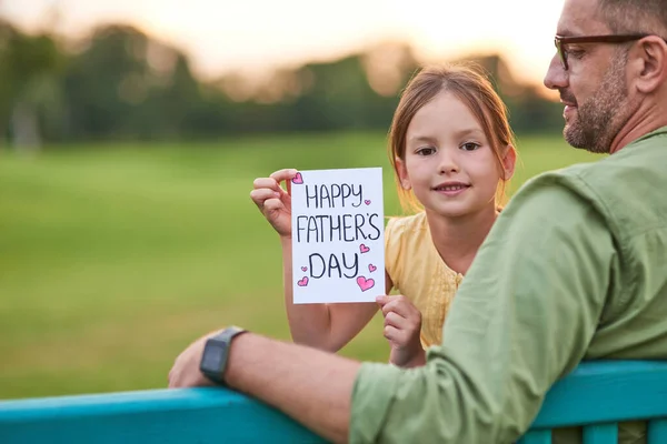 Linda niña mirando a la cámara y mostrando postal hecha a mano Feliz Día de los Padres mientras pasa tiempo junto con su padre en el hermoso parque verde — Foto de Stock