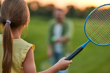 Bir yaz günü babasıyla parkta oynarken elinde badminton raketi tutan küçük bir kızın arka görüntüsü.