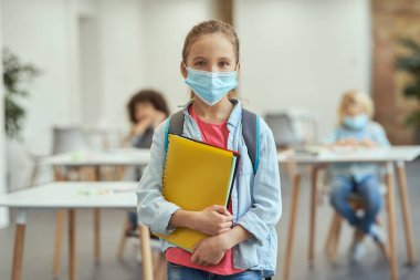 Covid19 'un yayılmasını önlemek için maske takan sevimli küçük okul kızının portresi. Kameraya bakıyor, sınıfta poz verirken ders kitaplarını tutuyor.