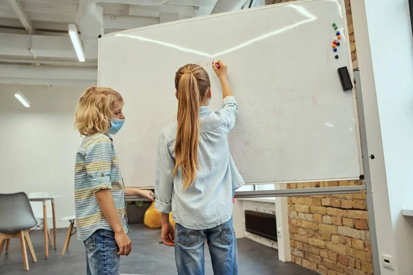 Małe dzieci w szkole, chłopiec i dziewczynka w masce ochronnej podczas pandemii koronawirusowej, piszący na pokładzie w klasie podstawowej. — Zdjęcie stockowe