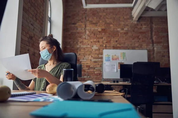 Diseñadora de interiores femenina con máscara protectora sosteniendo un plano mientras trabaja en un nuevo poject, sentada a la mesa en su oficina — Foto de Stock