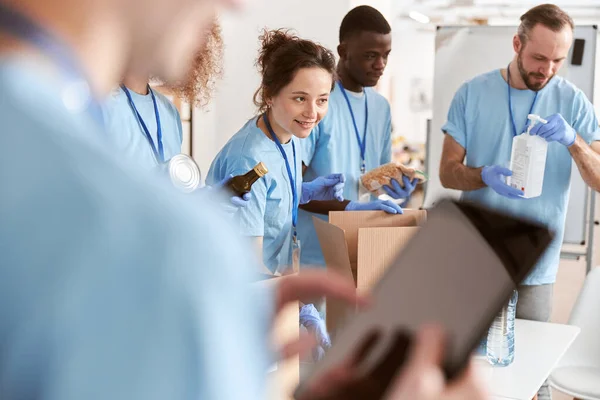 Sukarelawan pengalih perhatian dengan seragam biru menyortir bahan makanan dalam kotak kardus, bekerja sama dalam proyek donasi. Fokus pada wanita — Stok Foto