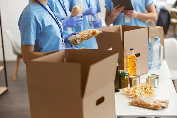 Gehakt schot van vrijwilligers in beschermende handschoenen sorteren, verpakken van voedsel in kartonnen dozen, werken aan donatie project binnen — Stockfoto