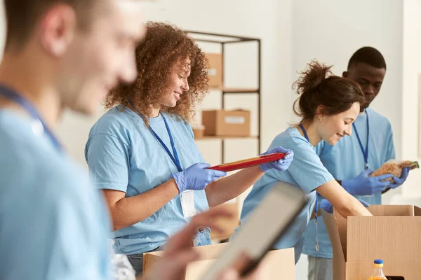 Seitenansicht einer glücklichen Freiwilligen, die lächelt, eine Packung Nudeln in der Hand hält, während sie Lebensmittel in Kartons sortiert und mit ihrem Team an einem Spendenprojekt arbeitet. Fokus auf die Frau — Stockfoto