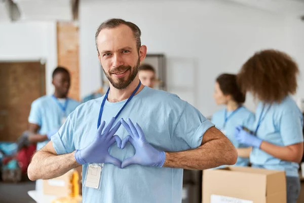 Retrato de un hombre caucásico voluntario en uniforme azul y guantes protectores mostrando el signo de amor del corazón, mirando a la cámara. Equipo de clasificación, embalaje de artículos en cajas de cartón — Foto de Stock