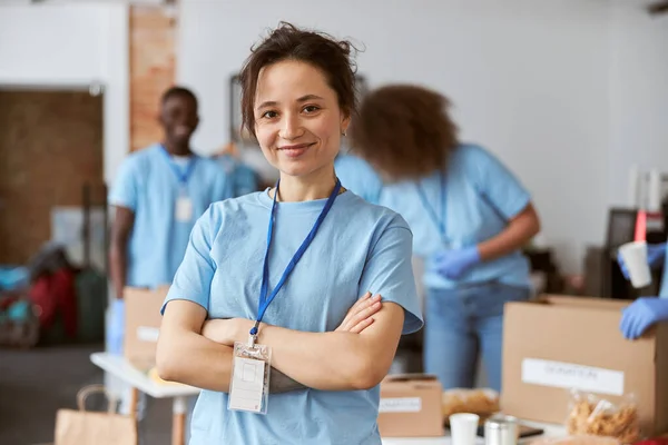 Retrato de una joven feliz, voluntaria en uniforme azul sonriendo a la cámara mientras está de pie con los brazos cruzados en el interior. Clasificación por equipos, artículos de embalaje en segundo plano — Foto de Stock