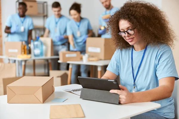 Attractive young woman, volunteer in blue uniform using tablet pc while sitting indoors, working on donation project. Team sorting, packing items in the background