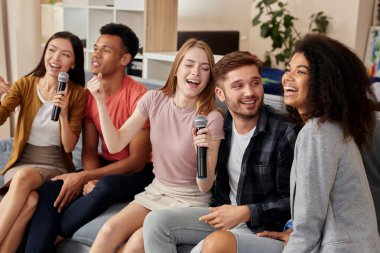 Müziğin tadını çıkar. Gülümseyen arkadaşlar evde karaoke yaparken heyecanlı görünüyorlar, mikrofonla şarkı söylüyorlar, modern dairede kanepede oturuyorlar.