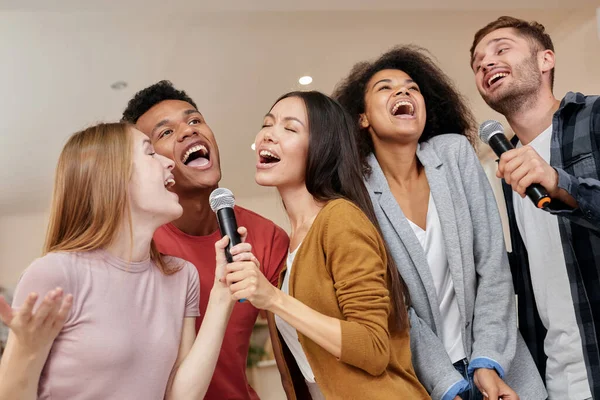 Bernyanyi untuk semua orang. Teman-teman yang ceria dan multikultural bernyanyi dengan menggunakan mikrofon saat bermain karaoke di rumah, berdiri di dalam ruangan — Stok Foto