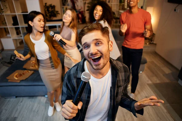 Tunjukkan kemampuan menyanyimu. Pria muda yang bahagia bernyanyi dengan mikrofon saat bermain karaoke dengan teman-teman di rumah, menari di apartemen modern — Stok Foto