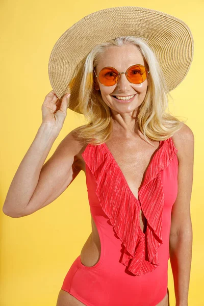 Studio photo de femme blonde heureuse en lunettes de soleil, maillot de bain rouge et chapeau de paille souriant à la caméra, ajustant son chapeau tout en posant, debout isolé sur fond jaune — Photo
