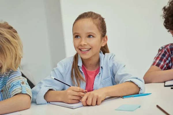 Härlig liten skolflicka ler bort, håller penna och lyssnar medan du studerar, sitter vid bordet i grundskolan klassrum — Stockfoto