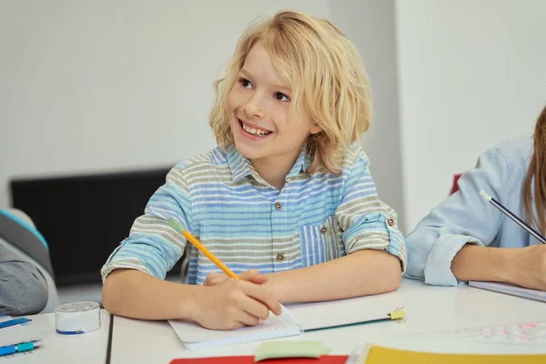 Boldog kis iskolás fiú vigyorog, ceruzával a kezében és hallgat, miközben tanul, ül az asztalnál az általános iskolai osztályteremben — Stock Fotó