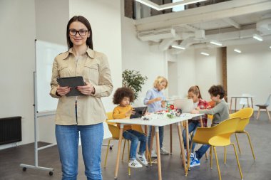Gündelik giyinen güzel bayan öğretmen kameraya gülümseyen ve sınıfta dururken tablet bilgisayar tutan. Arka planda robot üzerinde çalışan çocuklar