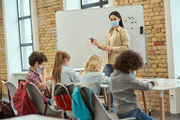Grupa dzieci w szkole podstawowej i nauczycielka nosząca maskę ochronną podczas pandemii koronawirusa pisząca zadania matematyczne na pokładzie w klasie — Zdjęcie stockowe