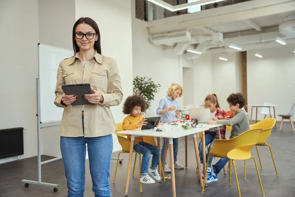 Hermosa maestra joven en ropa casual sonriendo a la cámara y sosteniendo la tableta PC mientras está de pie en un aula. Niños trabajando en robot bricolaje en el fondo —  Fotos de Stock