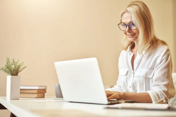 Perempuan dewasa yang bahagia memakai kacamata tersenyum sambil menggunakan laptop, bekerja di kantor — Stok Foto