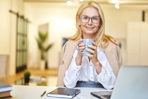 Wanita pengusaha cantik yang dewasa dengan kacamata tersenyum pada kamera dan minum kopi saat bekerja pada laptop di kantor — Stok Foto