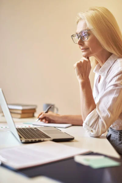Pengusaha wanita dewasa berkacamata tampak sibuk saat menggunakan laptop, membuat catatan, bekerja di kantor — Stok Foto