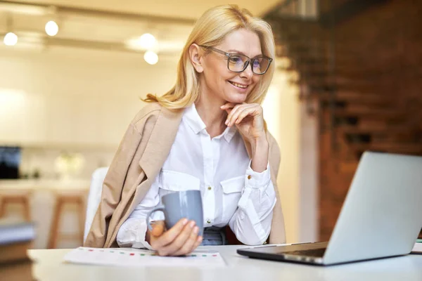 Senyum pengusaha dewasa berkacamata menggunakan laptop dan kopi sambil mengerjakan beberapa dokumen di kantor — Stok Foto