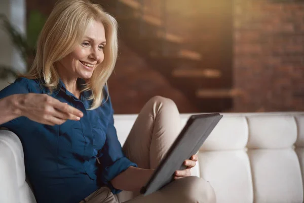 Wanita dewasa yang konten tersenyum saat menggunakan tablet digital pc, bersantai di sofa di rumah di ruang tamu — Stok Foto