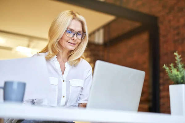 Pengusaha wanita dewasa yang sukses tersenyum sambil mengerjakan laptop sambil mengerjakan beberapa dokumen di kantor — Stok Foto