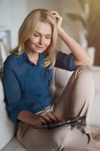 Wanita dewasa yang damai tersenyum, menggunakan tablet digital pc sambil bersantai di sofa di rumah di ruang tamu — Stok Foto