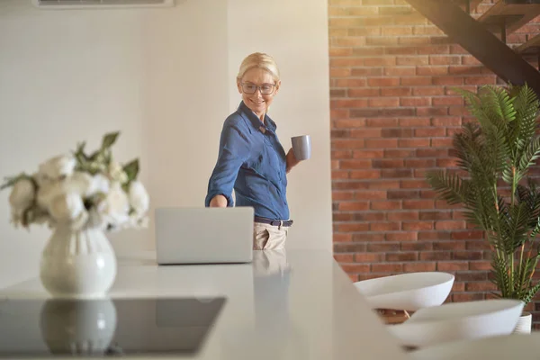 Neşeli sarışın, olgun bir kadın işe hazırlanırken dizüstü bilgisayar kullanıyor, modern mutfak odasında duruyor. — Stok fotoğraf