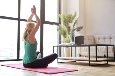 Sportif sarışın kadın yoga yapıyor, lotus pozisyonunda oturuyor ve evde dizüstü bilgisayar kullanarak online eğitim izliyor.