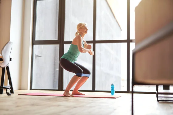 Seitenansicht einer fitten reifen Frau, die einen aktiven Lebensstil führt, zu Hause mit Widerstandsband trainiert, zu Hause vor großem Fenster auf einer Yogamatte steht — Stockfoto