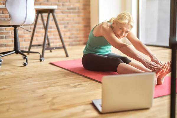 Flexible mature blonde woman in sportswear sitting on the floor, stretching her body while watching online video yoga class on a laptop at home — Stock Photo, Image
