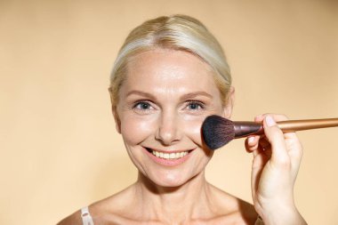 Bej arka planda izole edilmiş olgun sarışın kadın için doğal makyaj yapan yetenekli sanatçıya yakın çekim. Kameraya gülümseyen kadın, sonuçtan memnun.