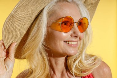 Güneş gözlüklü ve hasır şapkalı mutlu bir sarışının portresi, sarı arka planda yalnız poz verirken şapkasını düzeltiyor.