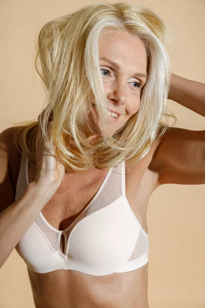 Studio portret van sensuele Kaukasische volwassen blonde vrouw model in witte lingerie glimlachen, aanpassen van haar haar terwijl poseren half naakt geïsoleerd over beige achtergrond — Stockfoto