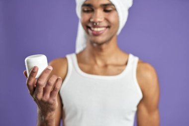 Mor arka planda elinde kozmetik yüz kremiyle gülümseyen genç adam yakın plan portresi