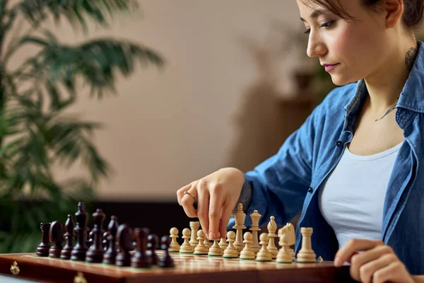 Молодая женщина смотрит на шахматные фигуры на доске и делает ход, играя в шахматы настольную игру дома в одиночку — стоковое фото