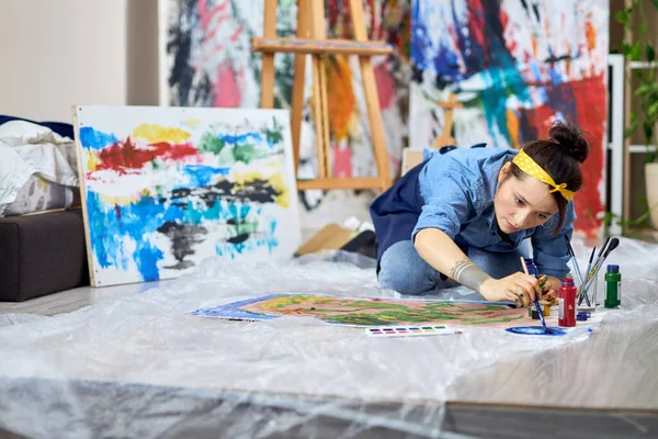 Getalenteerde vrouwelijke schilder in schort kiezen verf kleur, houden penseel tijdens het werken aan het schilderen, zitten op de vloer thuis workshop — Stockfoto