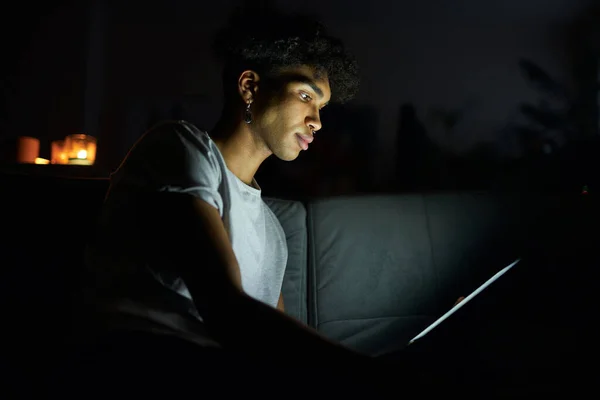 Seitenansicht eines fokussierten jungen Mannes, der nachts E-Books liest oder Videos mit dem Tablet-PC anschaut und zu Hause im dunklen Raum sitzt — Stockfoto