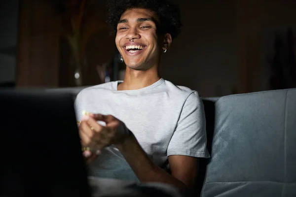 Fröhlicher junger Kerl lacht, während er auf einem Laptop einen Comedy-Film anschaut, und sitzt zu Hause auf einem Sofa im dunklen Raum — Stockfoto